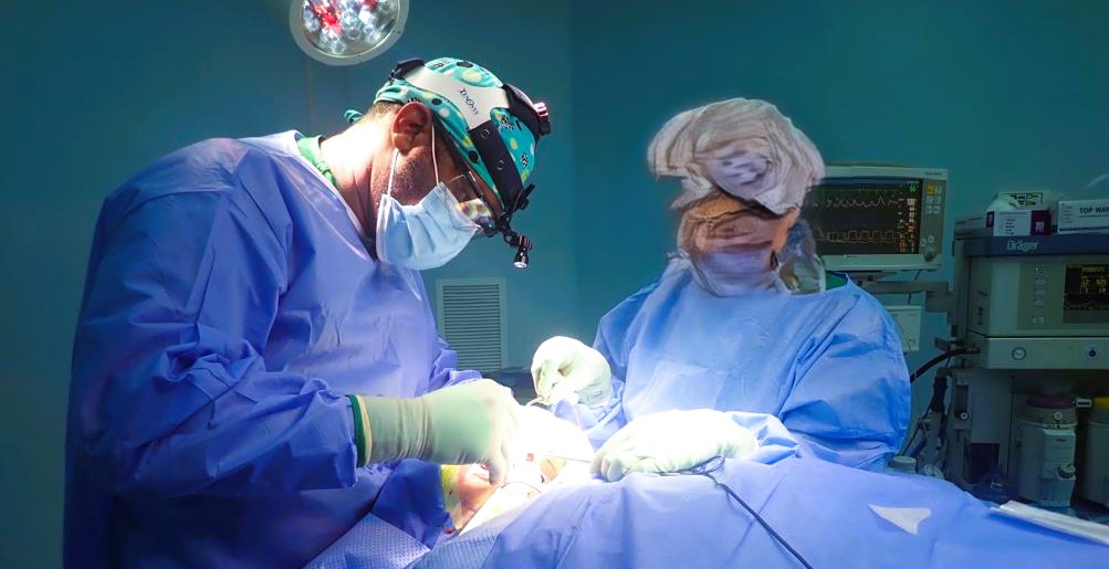 Surgeon wearing magnifying loupes and a headlight operating with an assistant beside anaesthesia equipment
