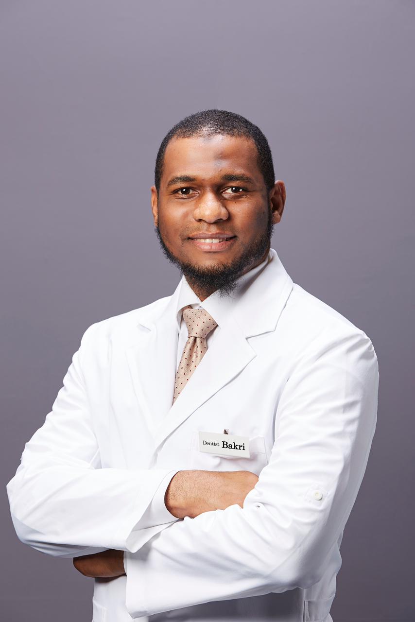 Studio portrait of Dr. Bakri in a white coat and tie with a name badge reading Dentist Bakri